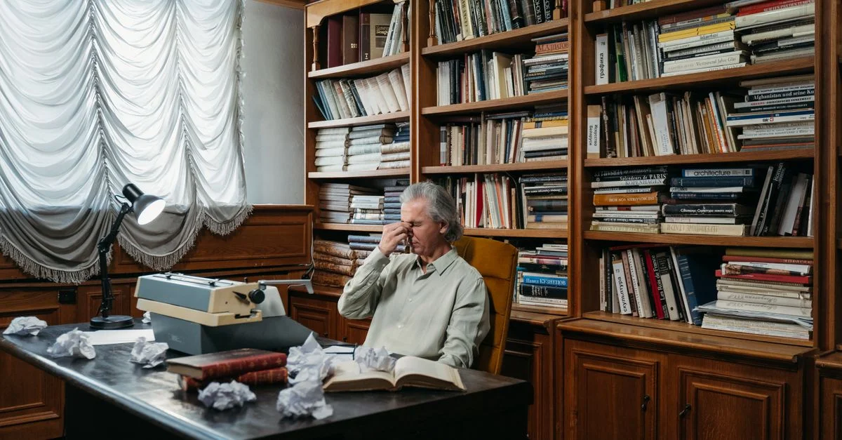 Homme senior fatigué à son bureau avec une machine à écrire et des livres dans un bureau classique.