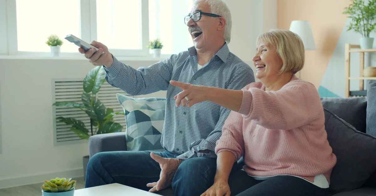 Un couple de personnes âgées rient et se détend sur le canapé en regardant la télévision à l'intérieur.