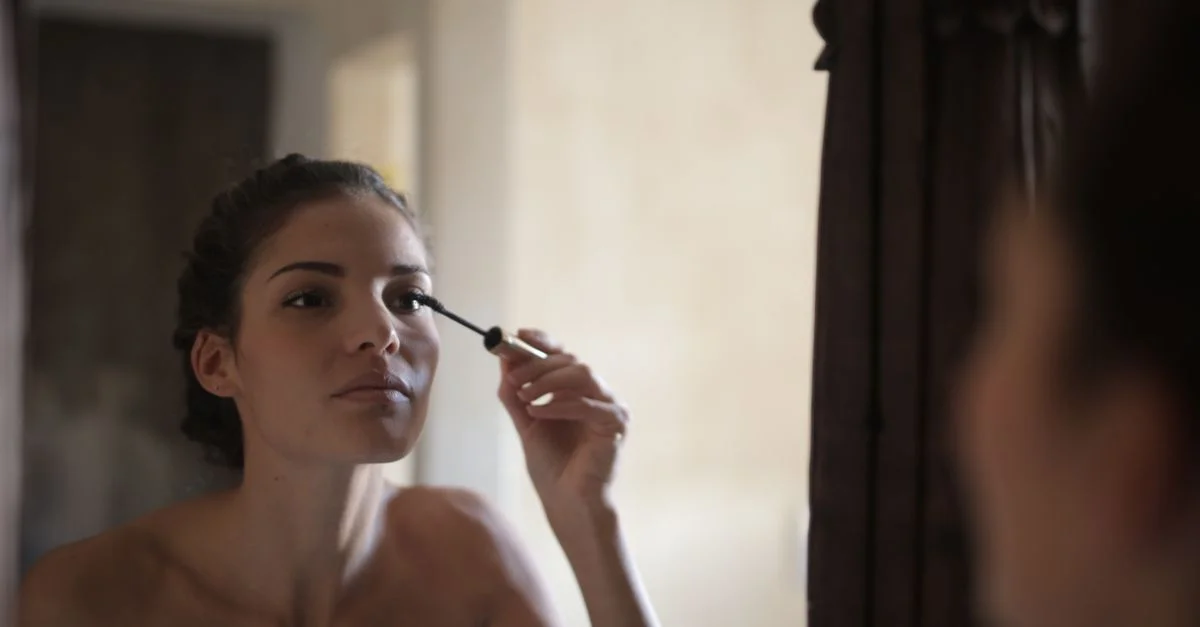 Jeune femme appliquant du mascara tout en se regardant dans un miroir.